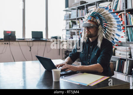 Man wearing Indian headdress and VR glasses in office, using laptop Stock Photo