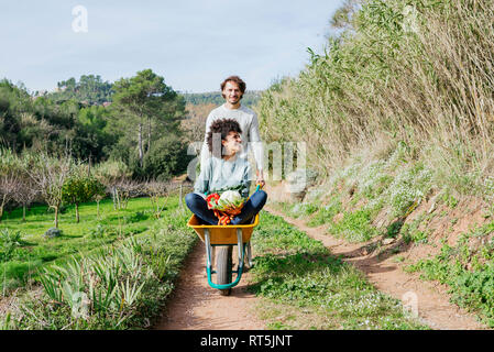 Woman sitting in wheelbarrow, holding fresh vegetables, man pushing her Stock Photo