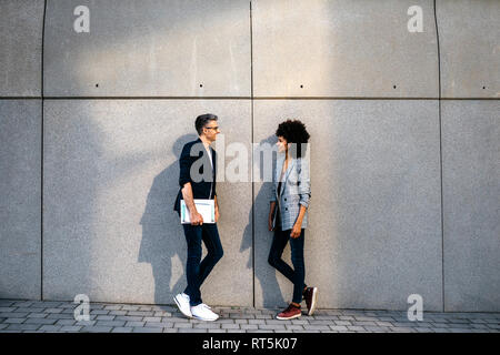 Two colleagues talking outdoors leaning against a wall Stock Photo