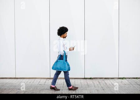 Man with bag walking in front of a white wall while looking at cell phone Stock Photo