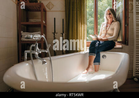 Woman sitting on window sill in the bathroom taking footbath while reading a book Stock Photo