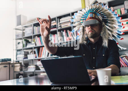 Man wearing Indian headdress and VR glasses in office, using laptop Stock Photo