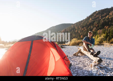 Mature man camping at riverside, sitting on tree trunk with map Stock Photo