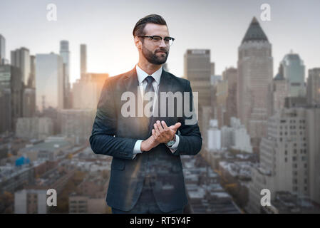 Satisfied with work done. Excited young businessman in suit rubbing his hands and looking away while standing against of morning cityscape background. Success concept. Business look Stock Photo