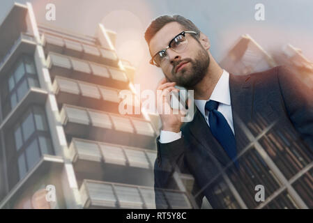 Business talk. Confident young businessman in suit talking on the mobile phone while walking outdoors with cityscape on the background. Digital. Internet. Communication. Stock Photo