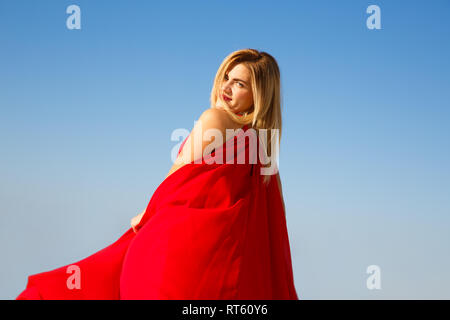 Blond woman in the red dress on the blue sky background. Stock Photo