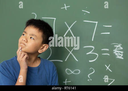 Thoughtful schoolboy doing math on green chalkboard in a classroom Stock Photo