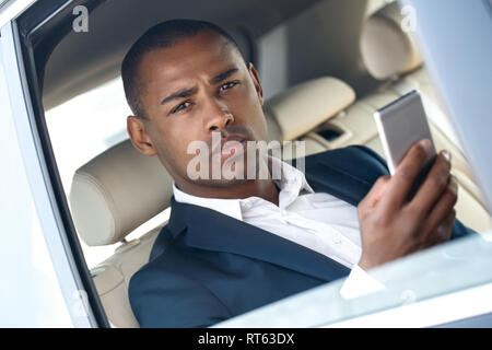 Young businessman sitting in car window opened browsing smartphone looking camera thoughtful close-up Stock Photo