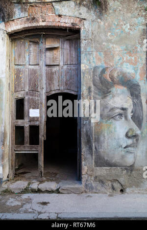 Entrance to an house with a broken door and walls an a poor state of repair with a mural painting of a woman's face on the right side. Havana, Cuba Stock Photo