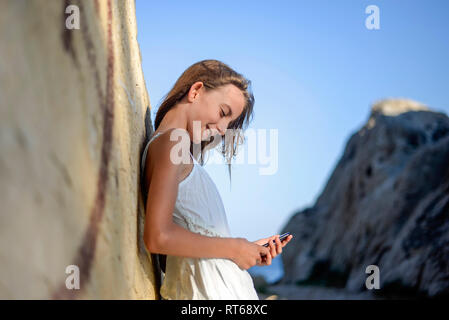 Croatia, Lokva Rogoznica, smiling girl leaning against rock looking at cell phone Stock Photo