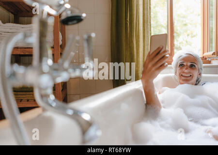 Portrait of happy woman in bathtub taking selfie with cell phone Stock Photo