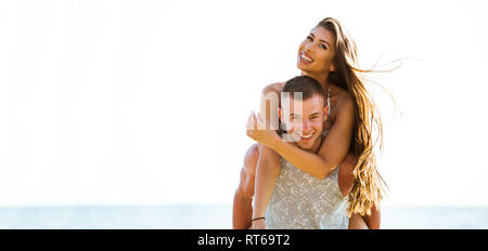 Young happy and joyful Caucasian adult romantic couple doing piggyback - Boyfriend carrying girlfriend on beach - with copy space Stock Photo