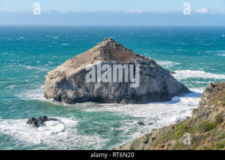 Waves crash against a large hat shaped sea stack isolated in the Pacific Ocean, just off the Big Sur shoreline in California. Stock Photo