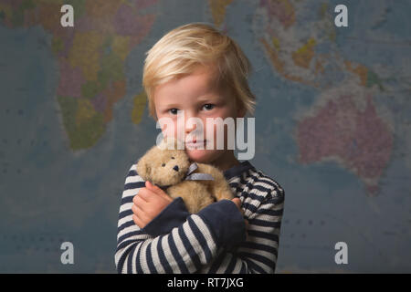 Cute blonde child cuddles his teddy bear in front of a large world map Stock Photo