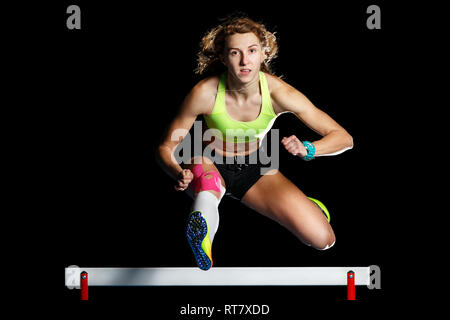 Young female athlete jumping over hurdle in sprint. Sprinter jumping over obstacle isolated on black background Stock Photo