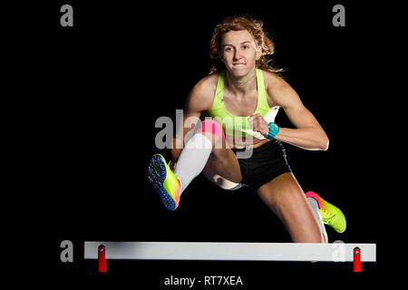 Young female athlete jumping over hurdle in sprint. Sprinter jumping over obstacle isolated on black background Stock Photo