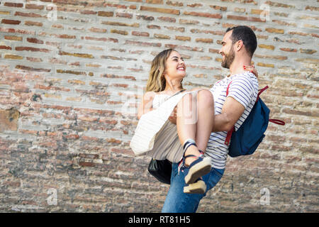 Happy man with backpack carrying girlfriend at brick wall Stock Photo