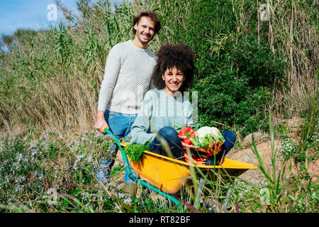 Woman sitting in wheelbarrow, holding fresh vegetables, man pushing her Stock Photo