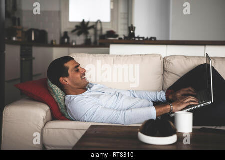 Smiling young man lying on sofa at home using laptop Stock Photo