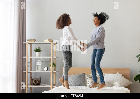Black mother and adolescent daughter holding hands jumping on bed Stock Photo