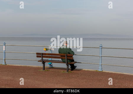 A lone man sat on a promenade bench looking out across Morecambe Bay next to a floral tribute Stock Photo