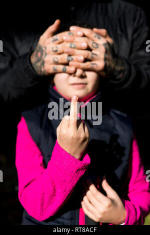 Girl giving the finger while father covering her eyes with his tatooed hands Stock Photo