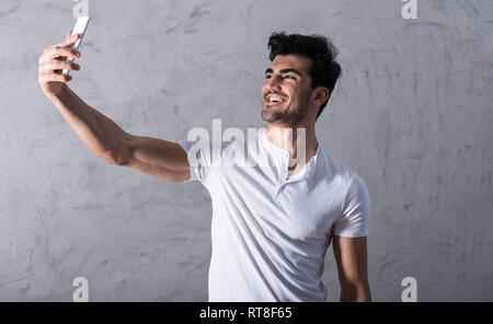 A happy handsome young man in a white tshirt taking a selfie with his smartphone in front of a grey wall in a studio. Stock Photo