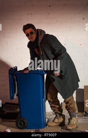 homeless man in dark glasses rummaging in blue trash container Stock Photo