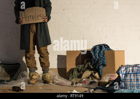 partial view of homeless man standing on garbage dump and holding piece of cardboard with "homeless" handwritten inscription Stock Photo