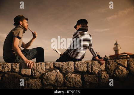 Side view of stylish guy in hat smoking pipe and elegant lady in cap reading volume and sitting on rocks near beacon and wonderful sky Stock Photo
