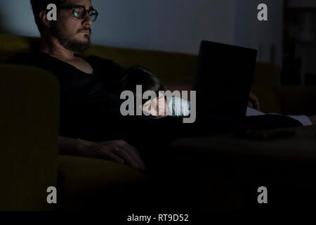 Father using laptop on couch at night with daughter sleeping Stock Photo