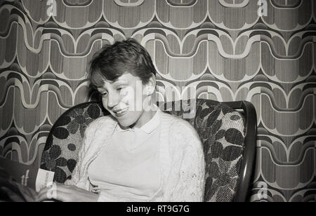 Late 1960s, historical, pretty teenage girl sitting on a chair, relaxing, reading a magazine in a room decorated with the whacky wallpaper of the era, England, UK. Stock Photo
