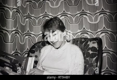 Late 1960s, historical, attractive, smiling girl sitting on a chair reading a magazine in a room with the patterned wallpaper of the era, England, UK. Stock Photo