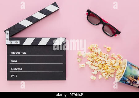 black clapperboard, glasses and a cup with scattered popcorn on a pink background Stock Photo