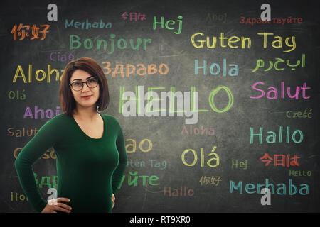Young woman student or teacher wearing glasses and holding hands on hips standing in front of blackboard written with the word hello in different lang Stock Photo