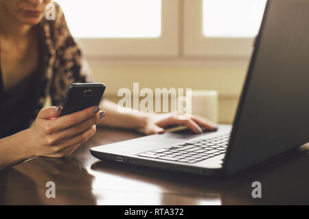 Unrecognizable young woman looking at smartphone while using laptop Stock Photo