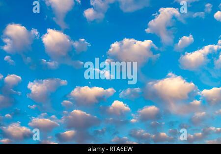Blue sky background with pretty fluffy white clouds tinged with color in evening light with side vignette in a full frame view Stock Photo