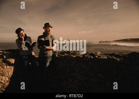 Stylish guy in hat and elegant lady in cap reading volumes near rocks on coast of sea and wonderful sky Stock Photo