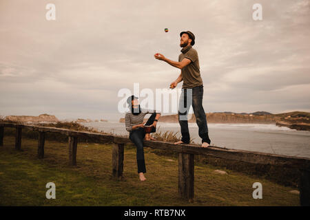 Young guy in hat juggling balls near elegant lady in cap with ethic drum sitting on seat near coast of sea and cloudy sky Stock Photo