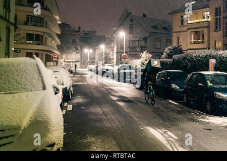 STRASBOURG, FRANCE - DEC 3 2017: Deliveroo delivery employee on bike in French city cycling fast for food delivery on time on a cold winter snowy night in residential neighborhood with cars parked Stock Photo