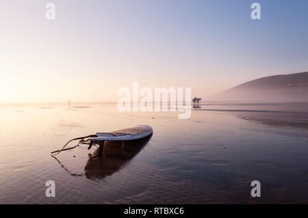 A surfboard on a beach relflected by the water on the sand. On a beautifully misty afternoon. Saunton, Devon, UK. Stock Photo