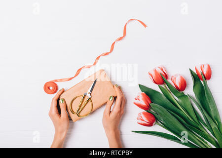View from above of female's hands holding gift box with golden scissors next to ribbon and bouquet of red tulips on white wooden table. Flat lay compo Stock Photo