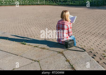 Blond girl in a red checkered shirt is sitting on a stone step and reading a book. Next to her is a black leather handbag. Bright, warm, spring day Stock Photo