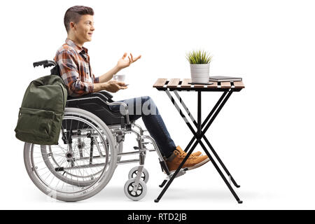 Full length profile shot of a disabled male student with a cup of coffee in a wheelchair sitting next to a wooden table isolated on white background Stock Photo