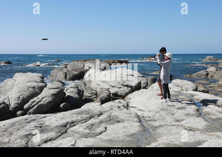 Man and woman fly and film a drone UAV over rocks and sea on Kaikoura Peninsula near the seal colony in the summer sun. New Zealand South Island Stock Photo