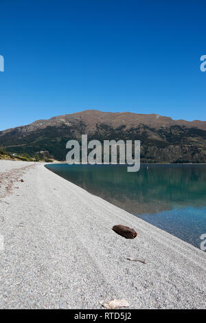 Beautiful Lake Hawea pebble beach with driftwood in summer blue sky clear water pebbles rocks New Zealand South Island Stock Photo