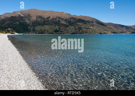 Beautiful Lake Hawea pebble beach with driftwood in summer blue sky clear water pebbles rocks New Zealand South Island Stock Photo