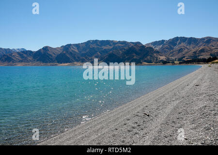 Beautiful Lake Hawea pebble beach with driftwood in summer blue sky clear water pebbles rocks New Zealand South Island Stock Photo