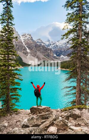 Hiker stands on the shore of a lake, stretches her arms into the air, mountain peaks at the back, turquoise glacial lake Stock Photo