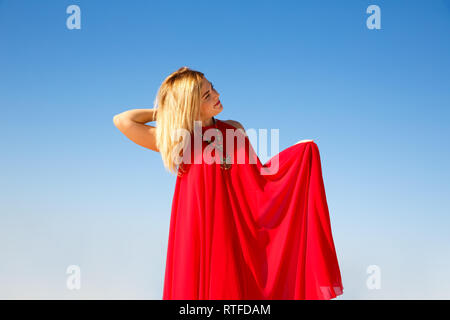 Blond woman in the red dress on the blue sky background. Stock Photo